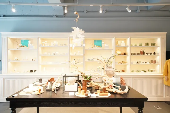 The interior of a shop with white shelving and display tables holding ceramics, wooden objects and dried branches, under track lighting.