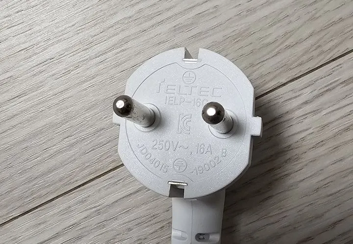 The moulded face of a white Korean plug with two round metal pins, a KC mark and clips on its edges, lying on a pale wooden floor. A rating is printed on its face.