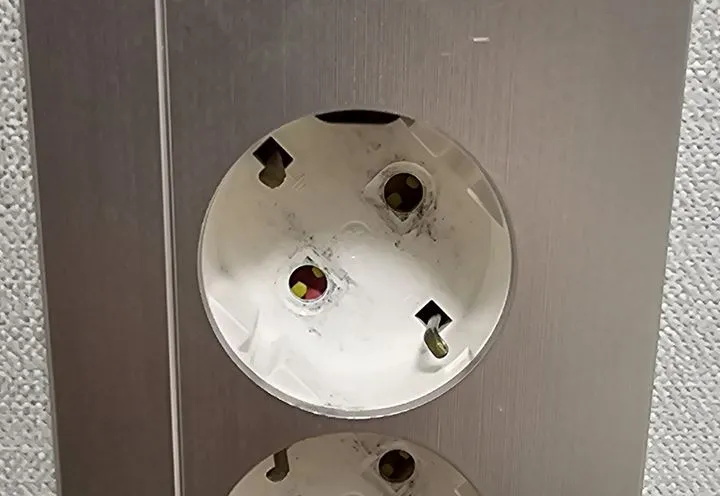 Close view of a silver-grey wall plate on a textured pale wall. A round white socket well sits set back from the plate, with two round pin holes and small earth contacts on the well wall; the top of a second well shows at the bottom edge.